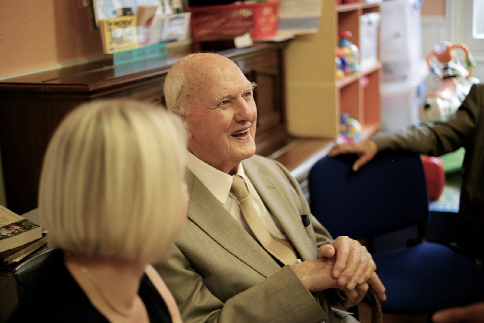 Man smiling at someone off camera man in brown suit jacket sitting beside woman in black long sleeve shirt